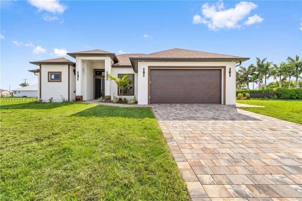 7 SW 19th Place, Cape Coral, FL 33991 Single-story exterior featuring a neutral stucco facade, multi-textured siding, a paved stone driveway, a deep-set covered entryway, and an attached two-car garage