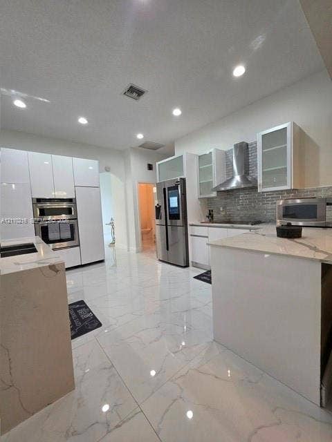 Kitchen featuring white cabinetry, stone-finish countertops, large-format tile flooring, a stainless steel range hood, and integrated wall ovens