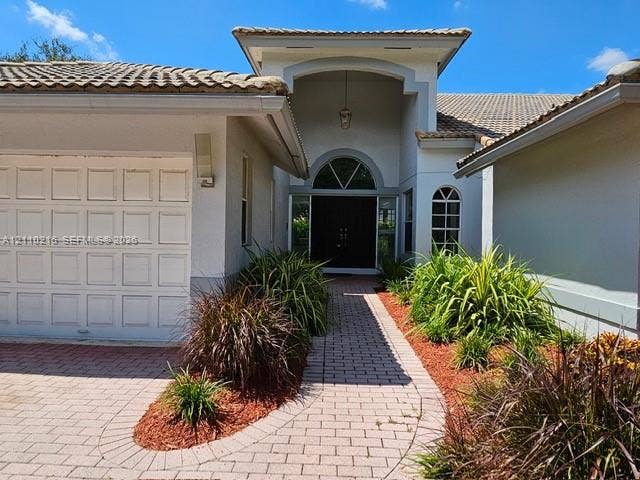 Stucco exterior featuring a dual-pitch tile roof, arched entryway with a double door, overhead transom window, and a paver stone walkway
