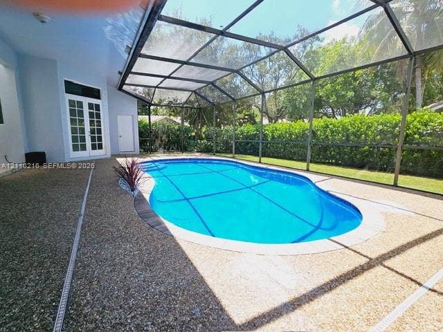 Screen-enclosed pool with a textured aggregate deck, surrounding privacy hedging, and multi-pane glass double doors