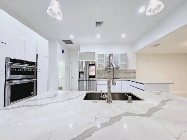 Kitchen featuring a large waterfall-edge island with stone-surface countertop, high-gloss flat-panel cabinetry, stainless steel double wall oven, stainless steel refrigerator, and undermount sink with a gooseneck faucet