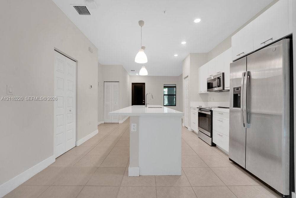 Kitchen featuring a central island with a solid-surface countertop, white flat-panel cabinetry, stainless steel appliances, recessed ceiling lighting, and large-format tile flooring