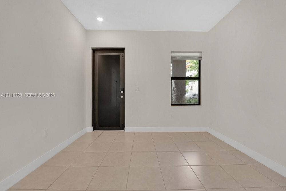 Neutral-toned interior room featuring large-format ceramic tile flooring, a dark-frame glass-panel door, a single-pane window with a roller shade, and recessed ceiling lighting
