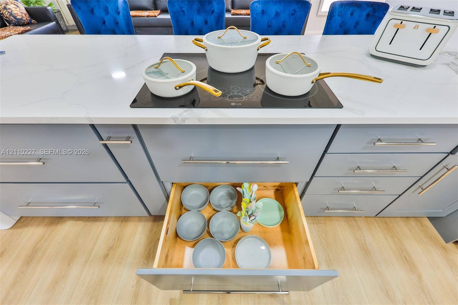 450 Bayshore Drive, Cape Coral, FL 33904 Kitchen island featuring a white surface with grey veining, a flush-mounted induction cooktop, light-grey flat-panel cabinetry with metal hardware, and deep-drawer storage with wood interiors, complemented by wood-finish flooring