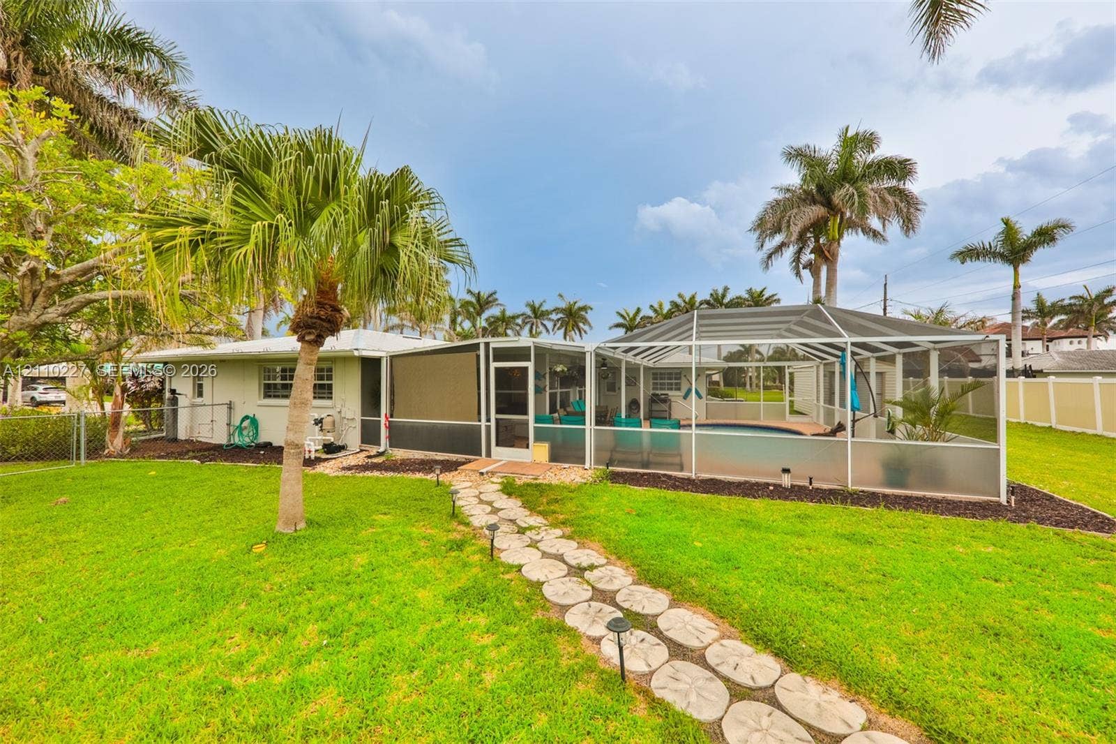 450 Bayshore Drive, Cape Coral, FL 33904 Expansive backyard featuring a manicured lawn, stone paver pathway, mature palm trees, and an enclosed swimming pool with a screened lanai