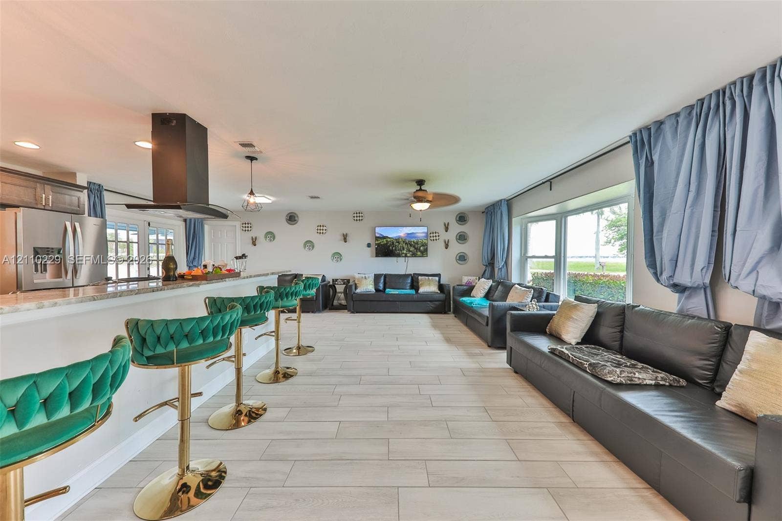 Open concept area featuring large-format ceramic tile flooring, an expansive breakfast bar with stone-finish countertops, stainless steel appliances, a modern black range hood, and floor-to-ceiling windows