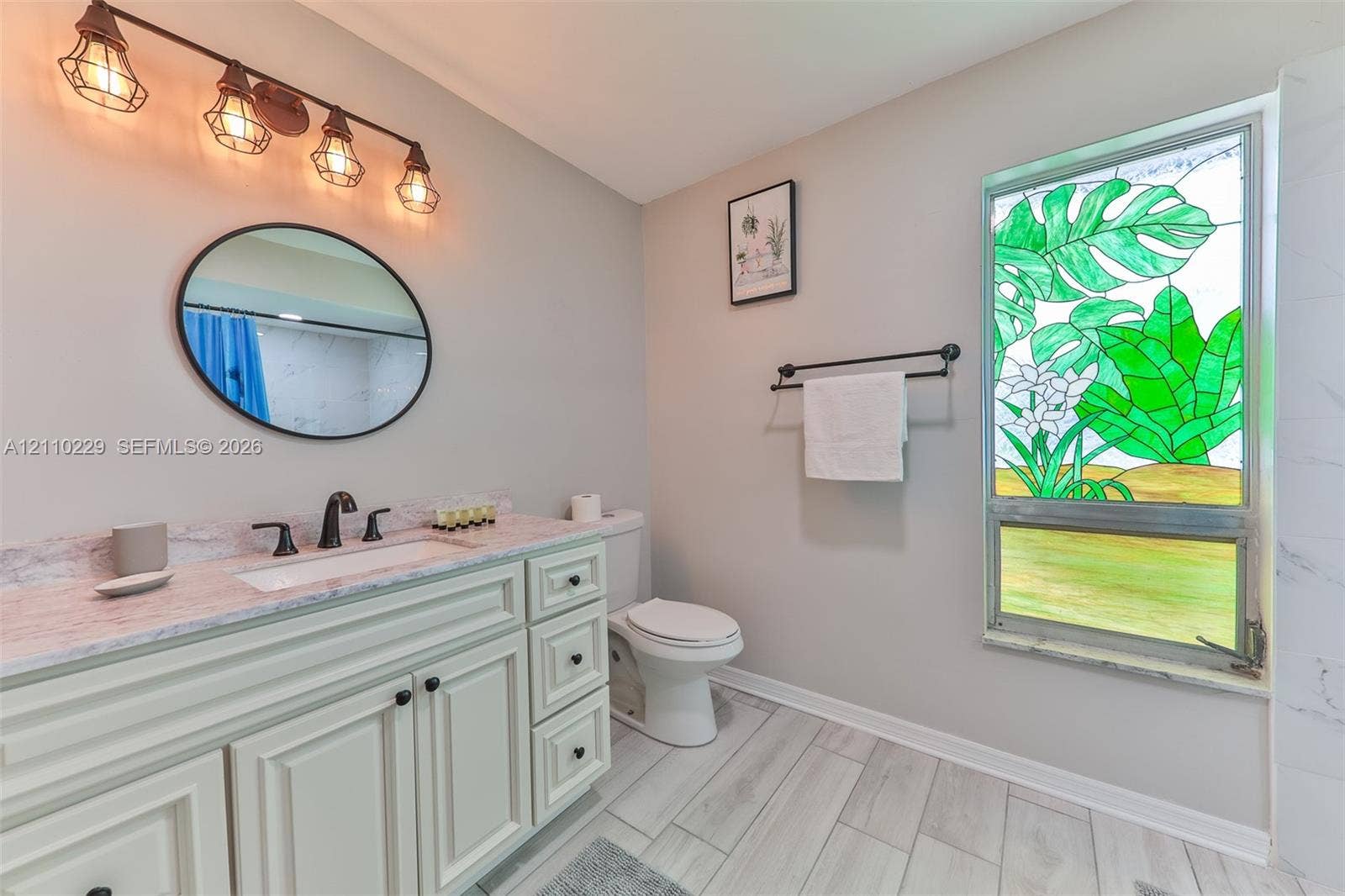 Bathroom featuring a custom stained-glass window, vanity with stone countertop, oval wall-mounted mirror, industrial-style four-light fixture, and wood-finish flooring