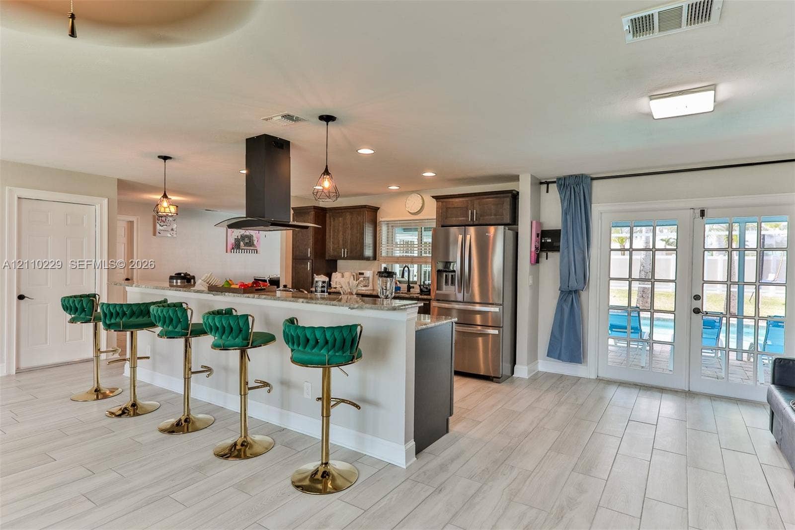Kitchen featuring wood-finish cabinetry, stone-surface breakfast bar, stainless steel refrigerator, matte black chimney-style range hood, and double glass-paned doors