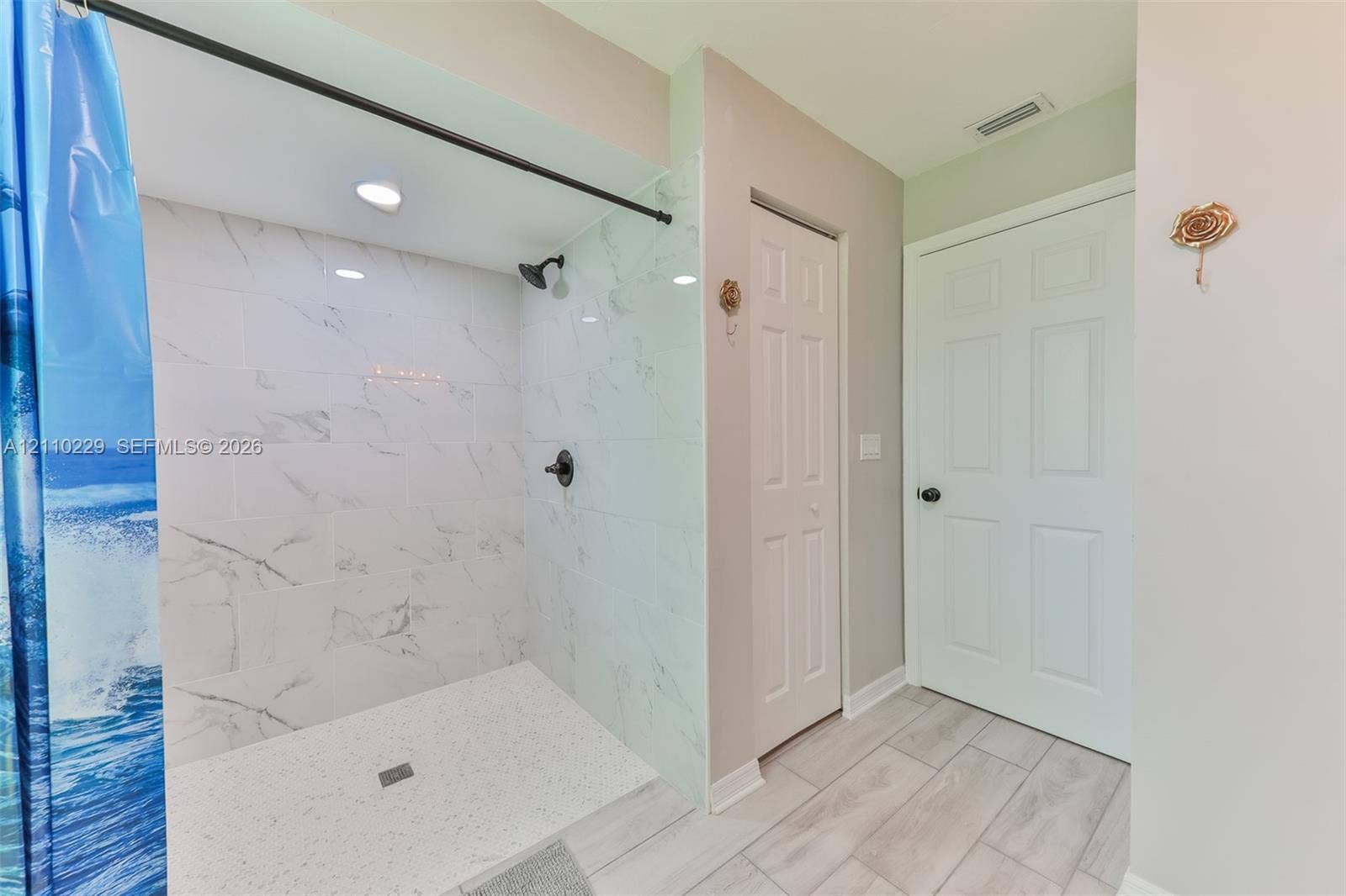 Walk-in shower featuring white stone-finish tile surround, dark metal hardware, recessed lighting, and wood-finish flooring