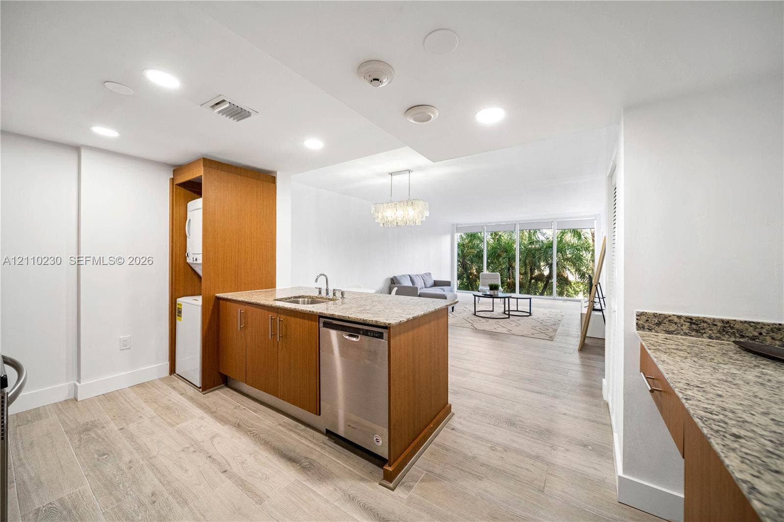 Kitchen featuring stone-finish countertops, stainless steel dishwasher, wood-finish cabinetry, wood-finish flooring, and a built-in laundry station with stacked unit