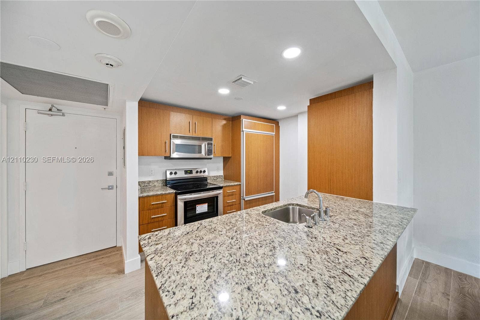 Kitchen featuring wood-finish cabinetry, stone countertops, a stainless steel undermount sink, built-in panel-ready refrigerator, and wood-finish flooring