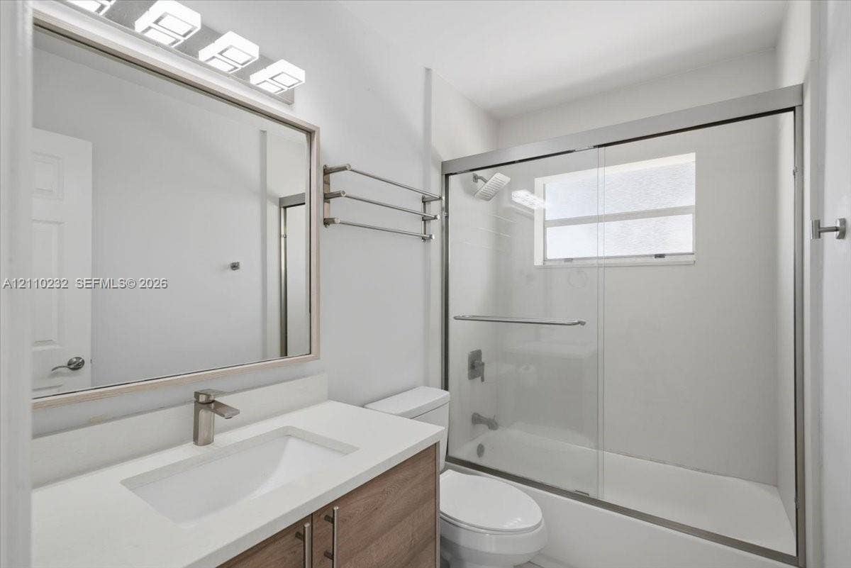 Wood-finish vanity with white countertop, integrated sink, brushed-metal hardware, sliding glass shower enclosure, and three-light wall-mounted fixture