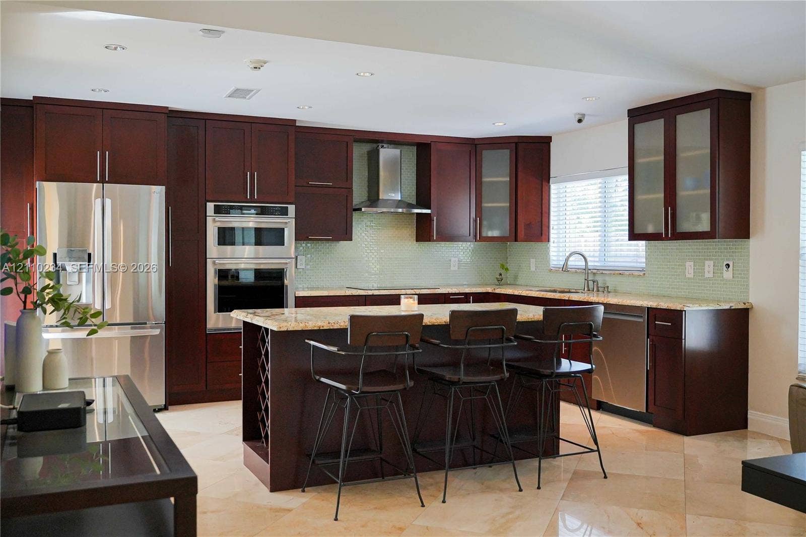 Kitchen featuring dark wood cabinetry, stainless steel appliances, a center island with built-in wine storage, stone countertops, and a glass tile backsplash