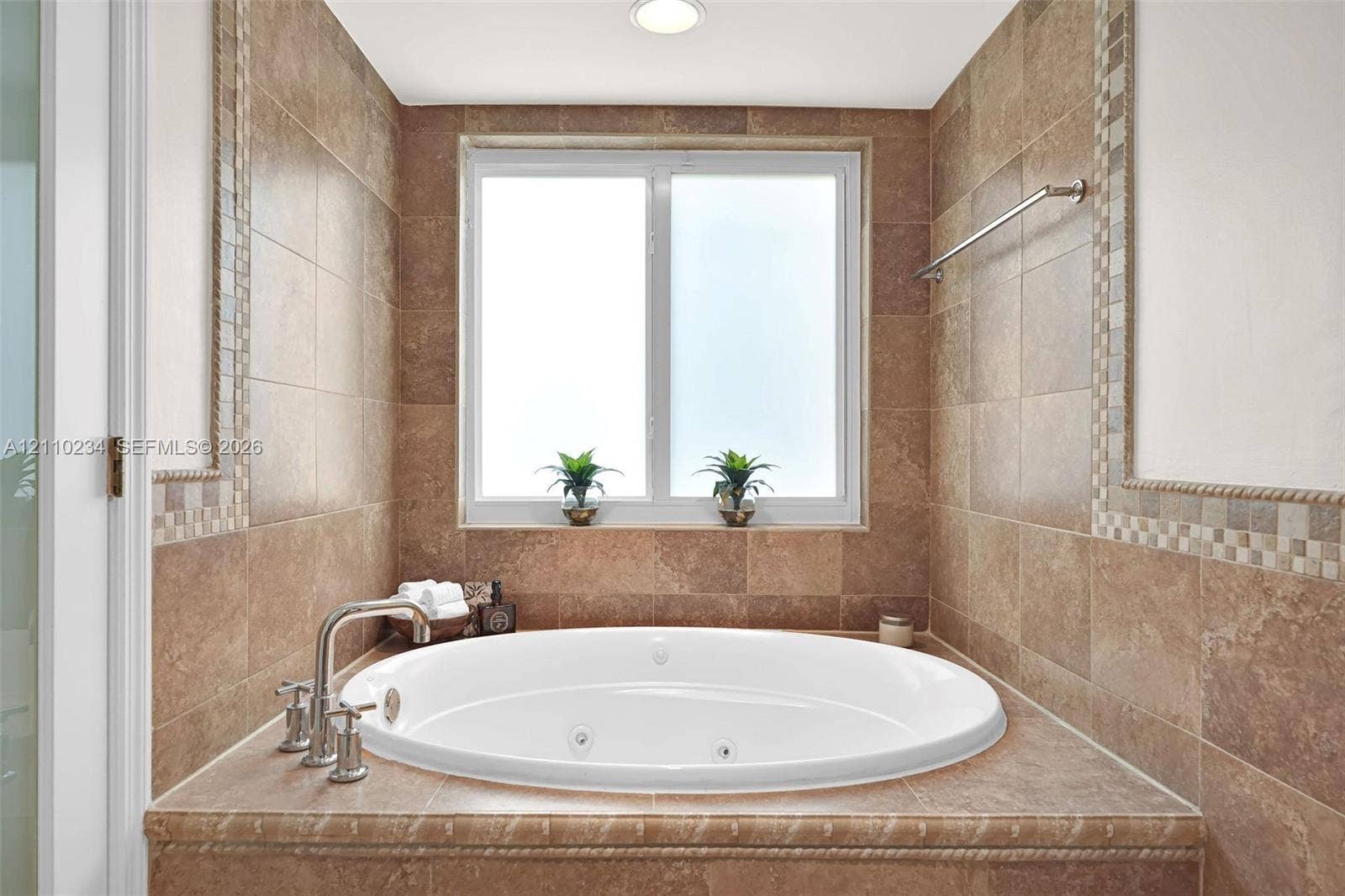 Oversized jetted soaking tub featuring a tile-surround deck, mosaic tile accents, polished chrome fixtures, and a dual-pane window