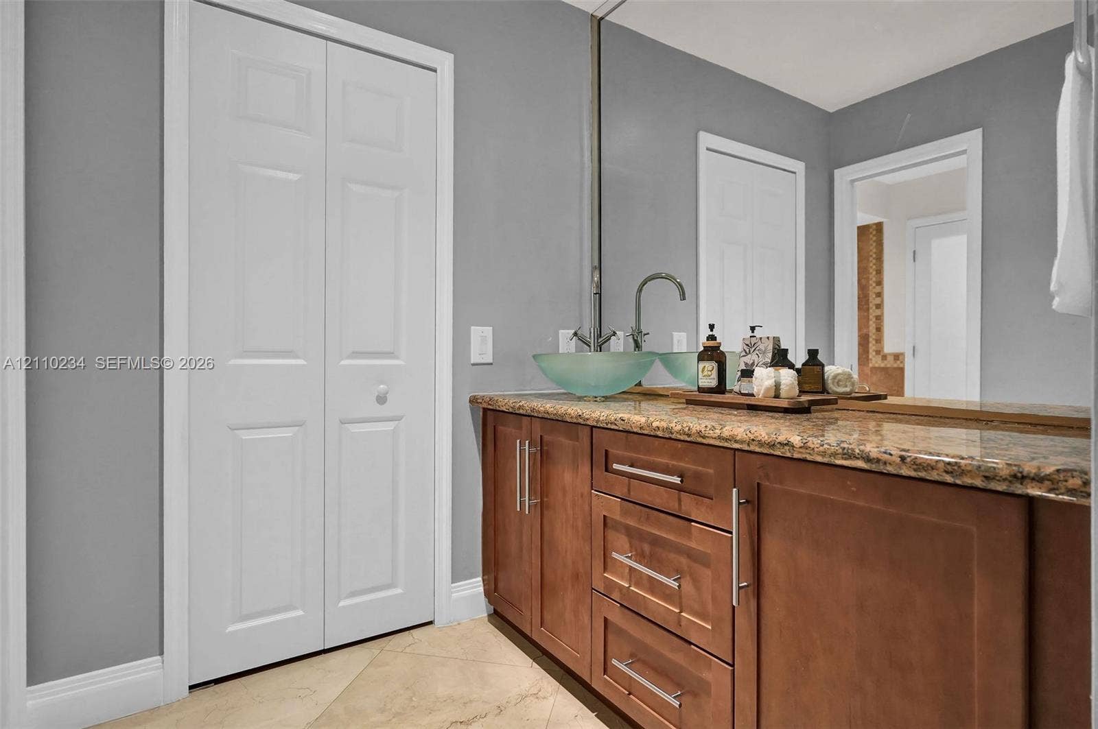 Bathroom vanity featuring a stone countertop, dual glass vessel sinks, high-arc faucets, wood-finish cabinetry with metallic hardware, and a large wall-mounted mirror
