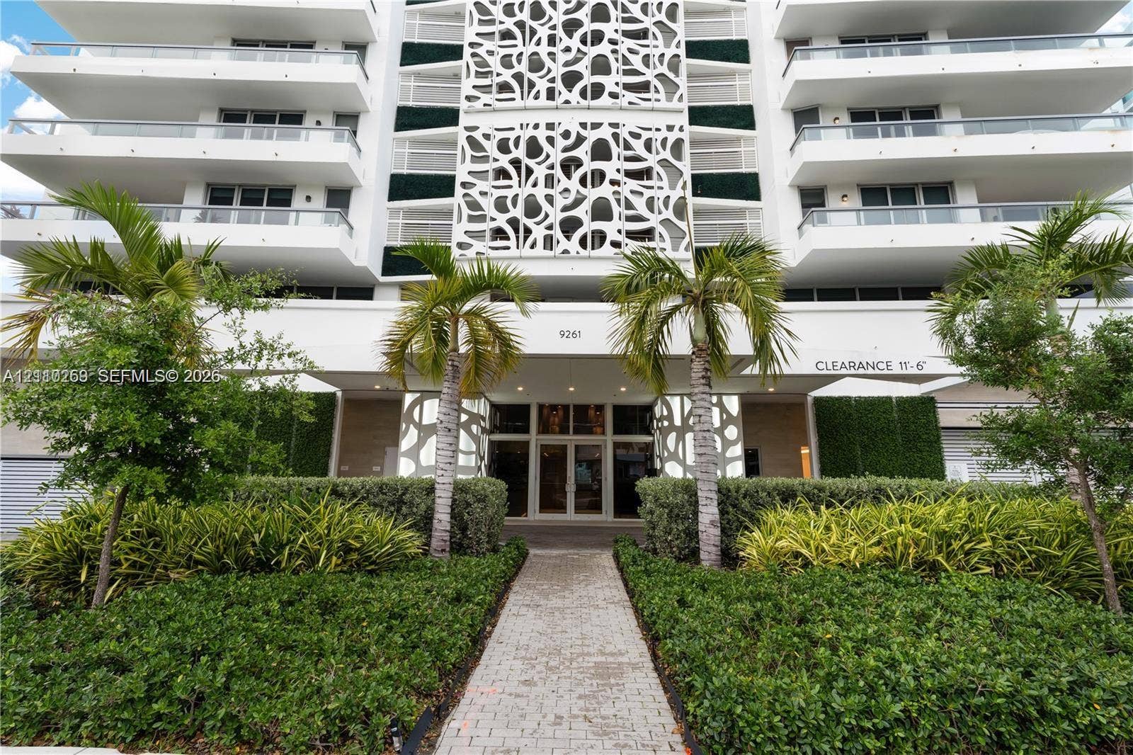 Modern mid-rise exterior featuring decorative latticework panels, tiered balconies with glass railings, landscaped hedging, and a paved stone entry path