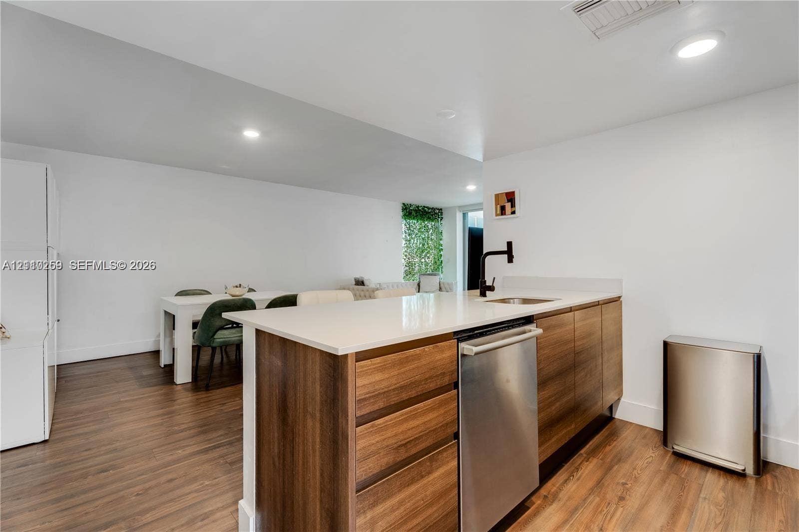 Kitchen island featuring wood-finish cabinetry, a white countertop, an undermount sink with a matte black faucet, and an integrated stainless steel dishwasher, complemented by wood-finish flooring and recessed lighting