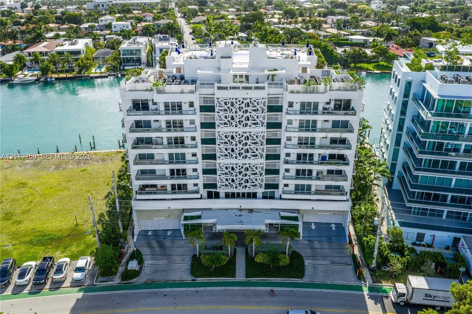 Waterfront high-rise building featuring decorative facade screening, private balcony levels, ground-level covered parking, and palm tree landscaping