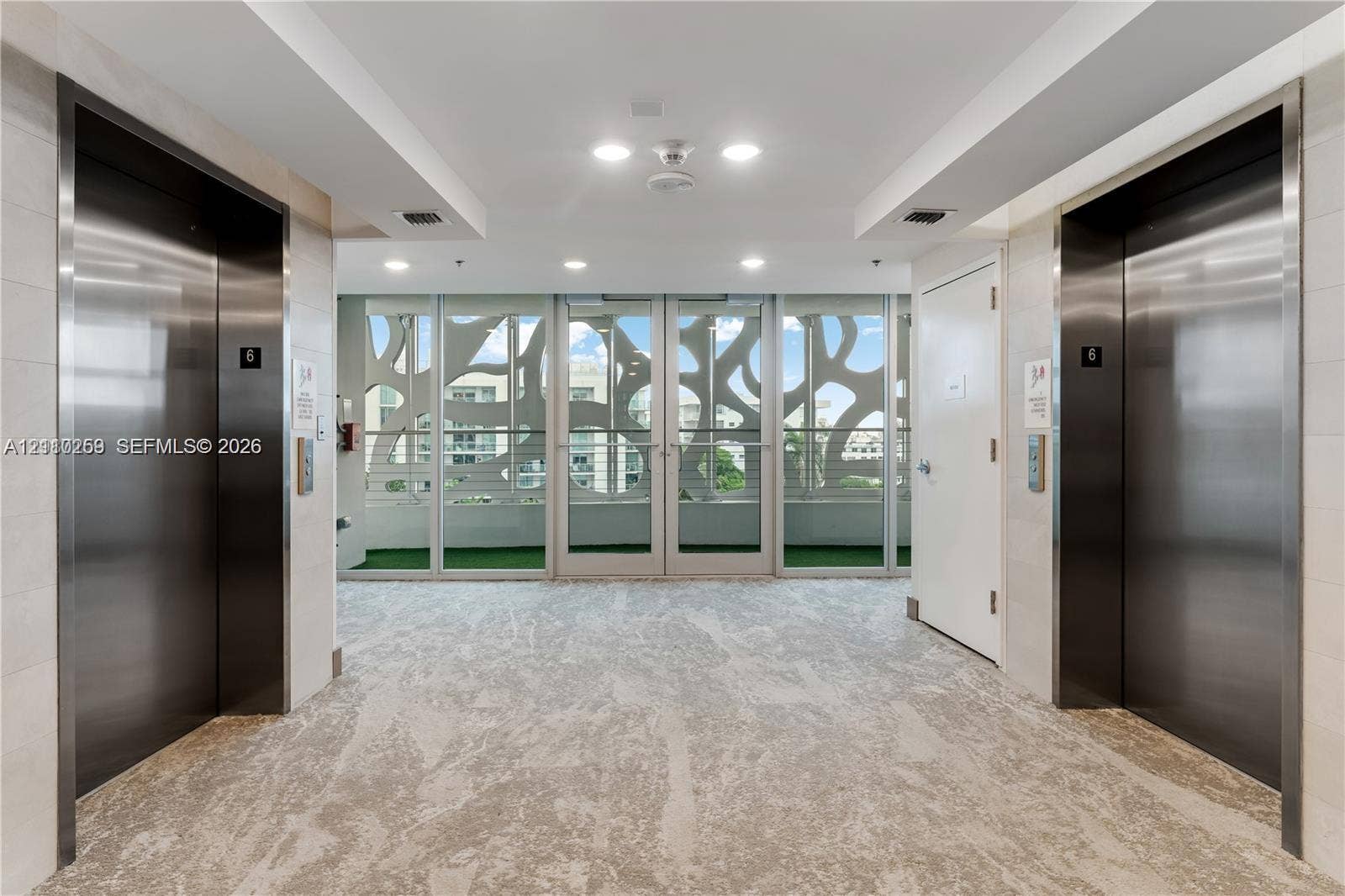 Elevator lobby featuring stainless steel doors, neutral patterned carpeting, recessed lighting, and floor-to-ceiling glass paneling with decorative metal screening