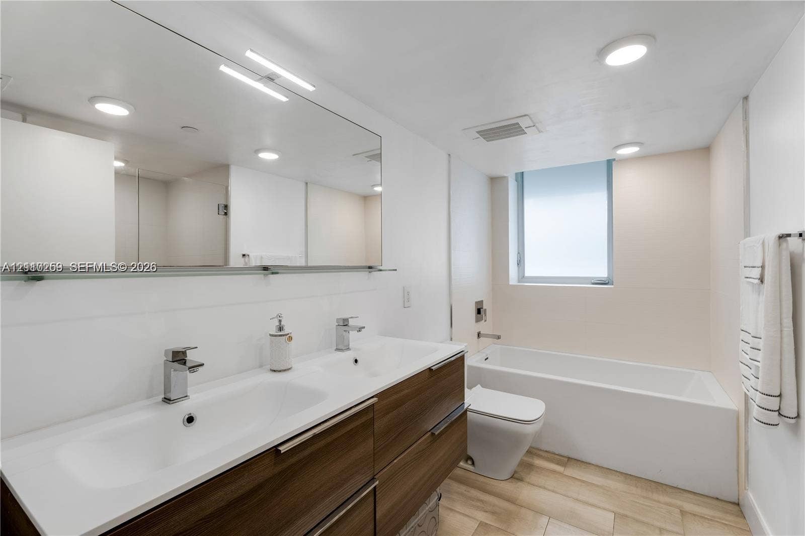 Double vanity with wood-finish cabinetry, integrated rectangular sinks, polished chrome faucets, and large wall-mounted mirror, complemented by wood-finish flooring and a white soaking tub