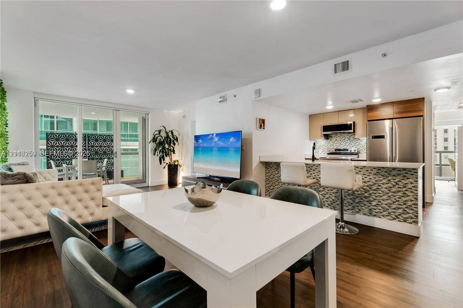 Open-concept living space featuring wood-finish flooring, recessed lighting, a kitchen island with stone-tiled base, integrated stainless steel appliances, and sliding glass balcony doors