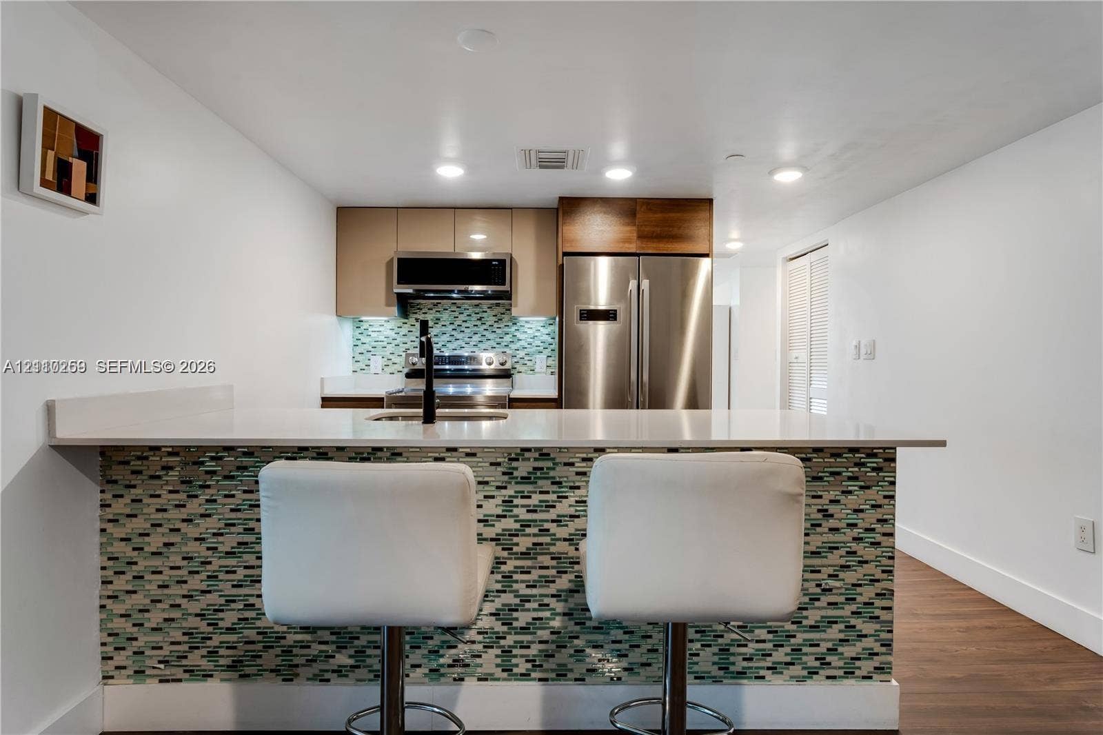 Kitchen featuring high-gloss cabinetry, stainless steel appliances, a multi-tone glass tile backsplash, and a white countertop with an integrated breakfast bar