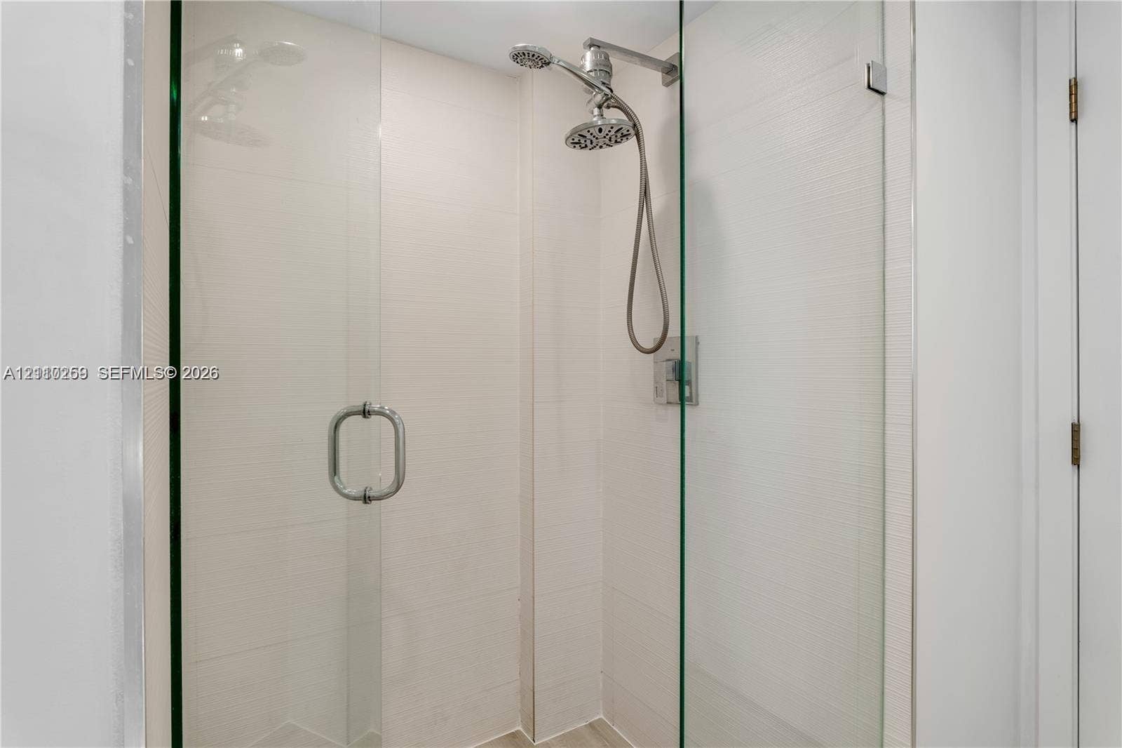 Walk-in shower featuring a frameless glass enclosure, textured wall tile, and an adjustable handheld showerhead with a polished metal finish