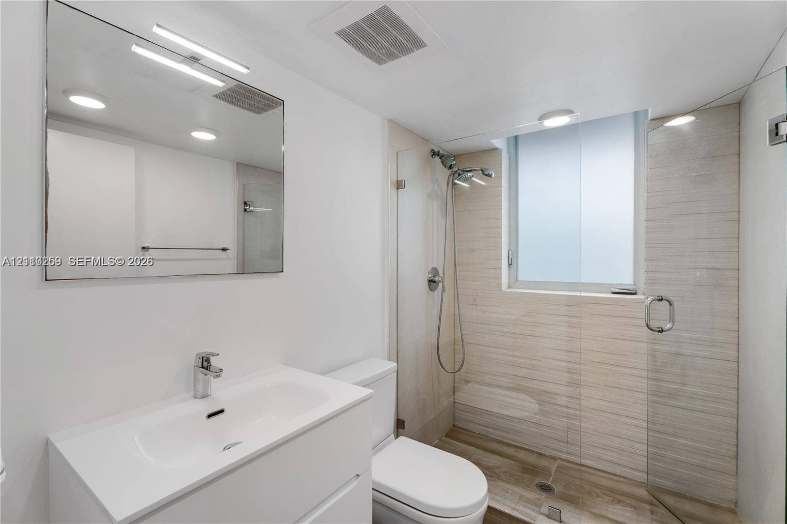 Modern bathroom featuring a floating white vanity, frameless glass shower enclosure, integrated wall-mounted lighting, and neutral stone-finish tile work