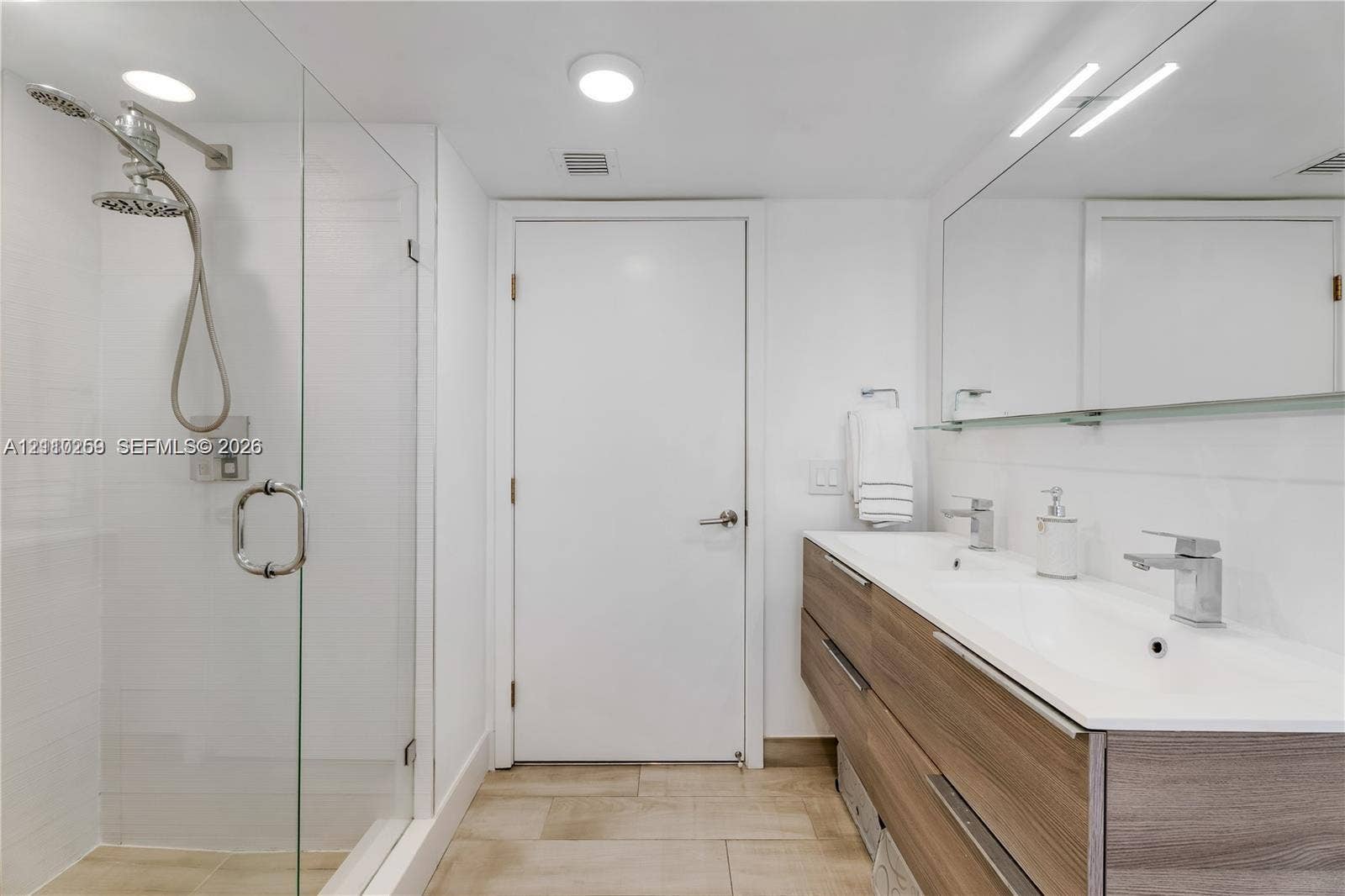 Floating wood-finish vanity with a double integrated sink, wall-spanning mirror, glass-enclosed shower with a handheld fixture, and neutral tile flooring