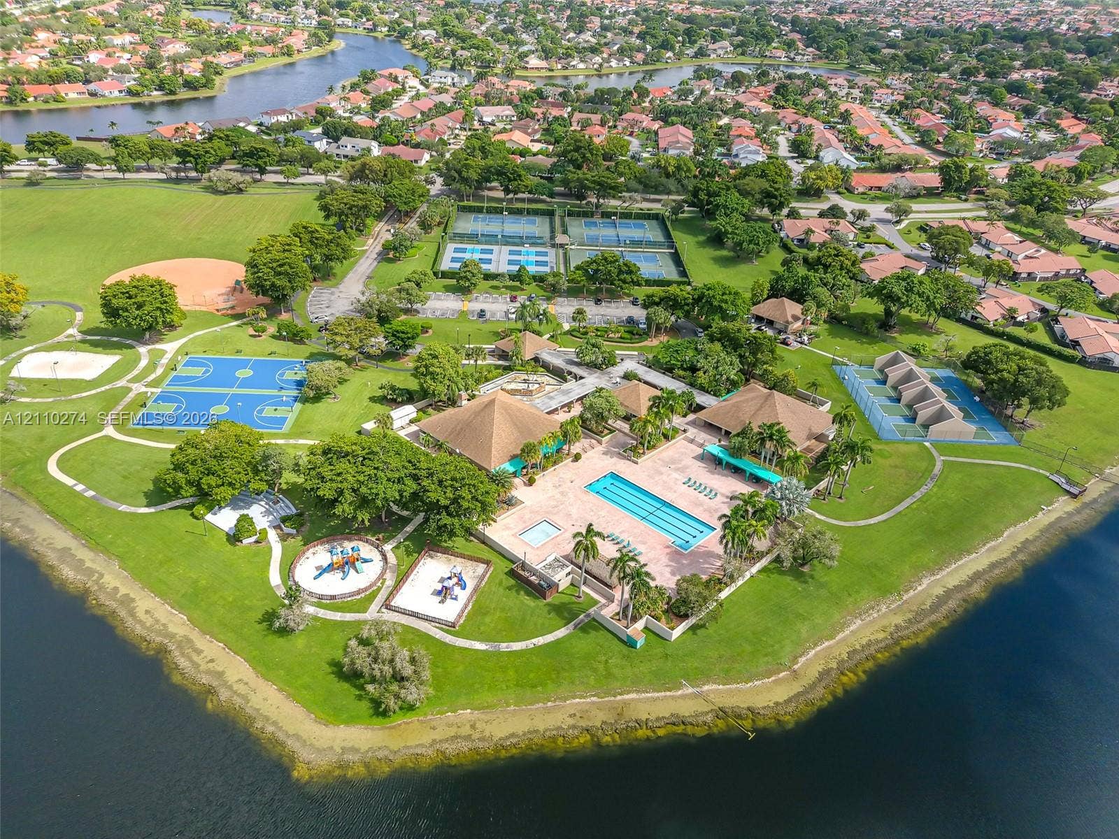 14955 SW 49th Lane, #5E, Miami, FL 33185 Recreational facility featuring a swimming pool, multiple tennis courts, multi-purpose sports courts, paved walking paths, and mature landscaping situated along a waterfront perimeter