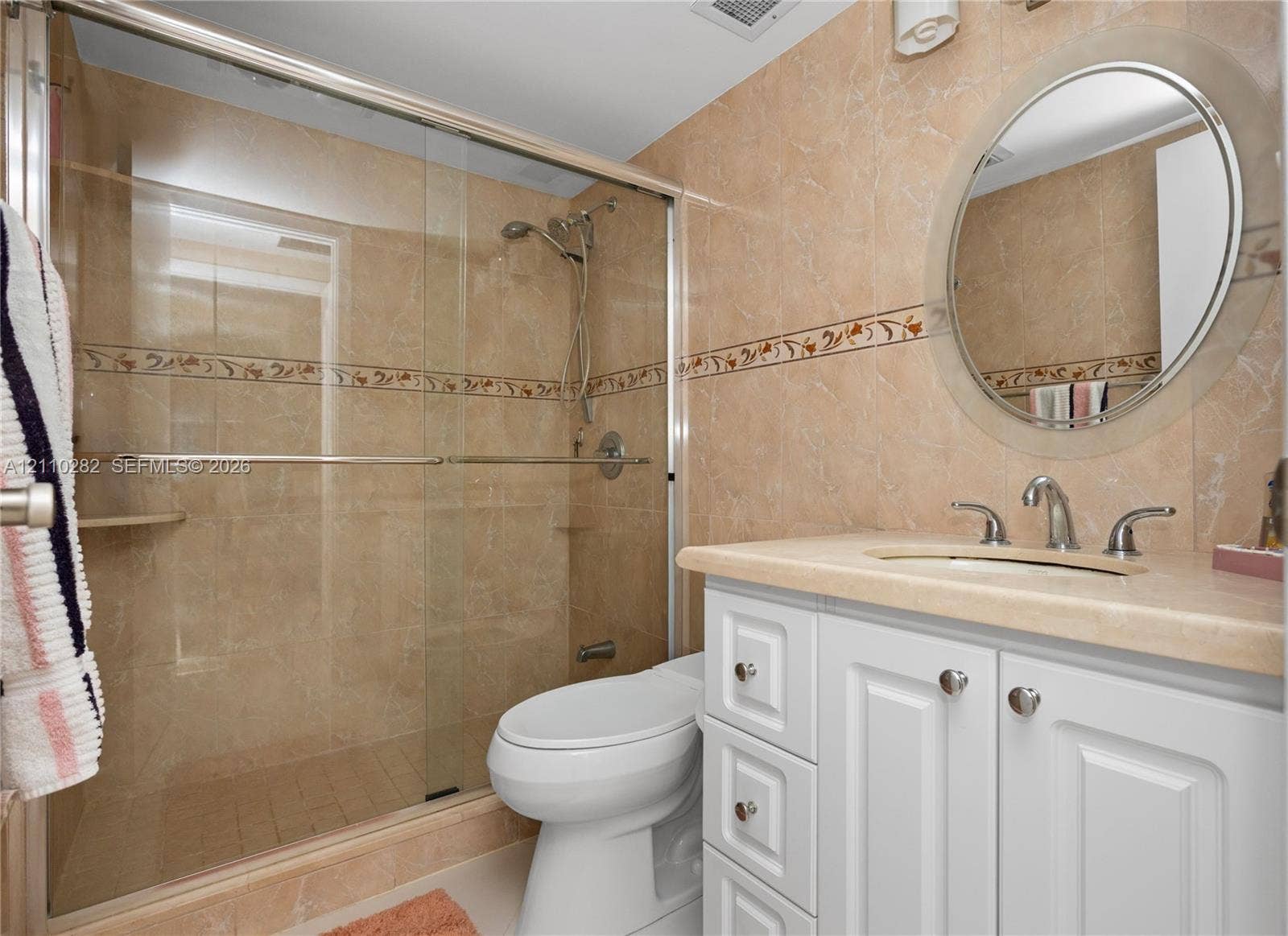 Bathroom featuring a white vanity with a neutral countertop, an integrated sink, a circular wall-mounted mirror, and a walk-in shower enclosure with tile surround and decorative accent border