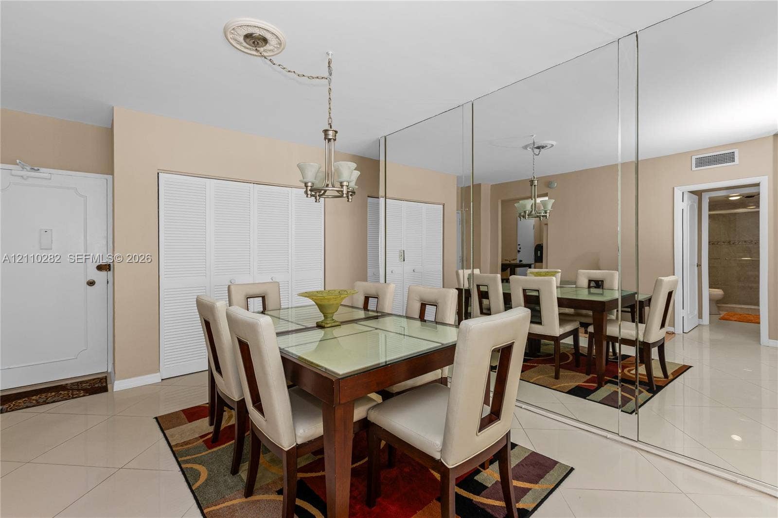 Large-format light-colored tile flooring, full-wall reflective mirror paneling, ceiling-mounted multi-light fixture with decorative medallion, and built-in closet storage with louvered doors