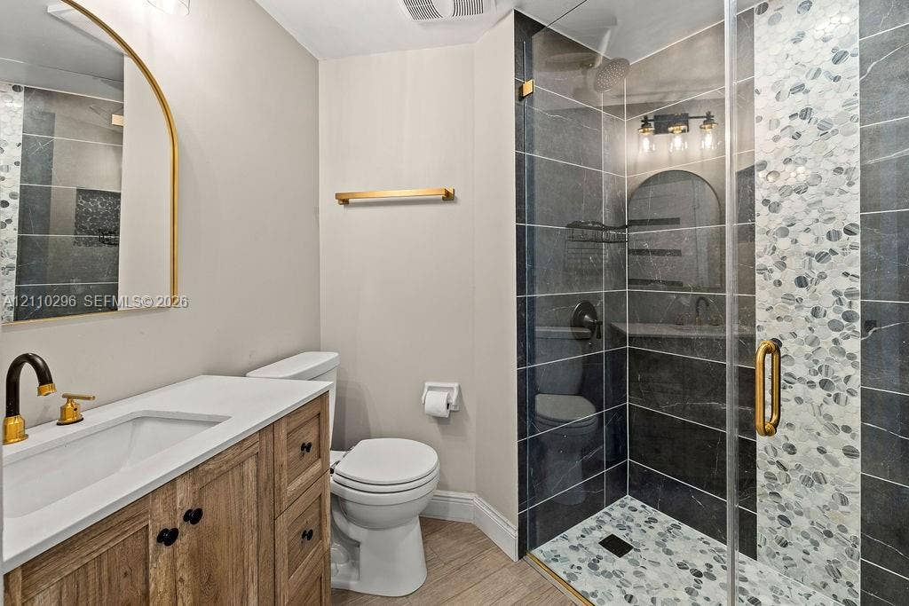 Bathroom featuring a wood-finish vanity with a stone countertop, an arched gold-rimmed mirror, a walk-in shower with stone tile accents, and gold hardware finishes