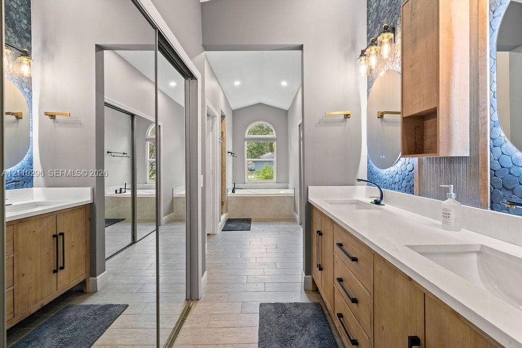 Dual wooden vanities with white countertops, integrated sink basins, textured blue tile accent walls, and floor-to-ceiling mirrored closet doors, featuring tile flooring and a soaking tub framed by an arched window