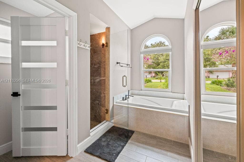 Bathroom featuring an arched-window soaking tub, glass-enclosed walk-in shower with tile surround, wood-finish tile flooring, and a multi-panel door with frosted glass inserts
