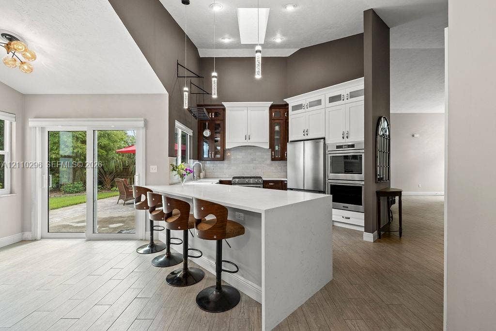Kitchen featuring an expansive waterfall-edge quartz island, stainless steel double wall ovens, glass-paneled upper cabinetry, tile flooring, and a glass sliding door