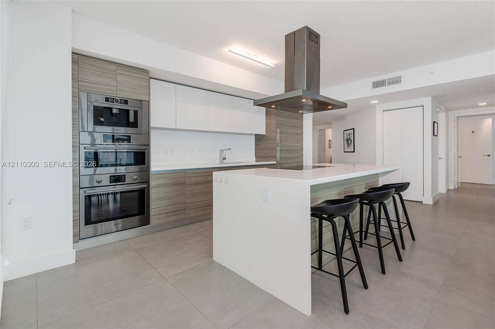 Contemporary kitchen featuring flat-panel cabinetry with wood-finish and solid white fronts, integrated stainless steel double wall ovens, a center island with a white waterfall countertop, and a stainless steel chimney-style range hood