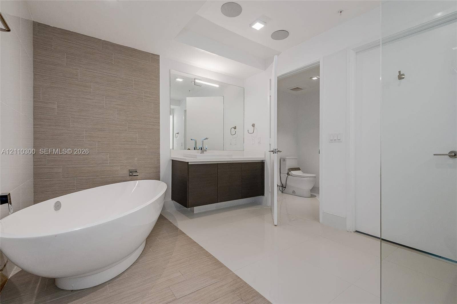 Master Bathroom Freestanding oval soaking tub, wall-mounted faucet, floating wood-finish vanity with a rectangular sink, large format tile flooring, and textured tile accent wall