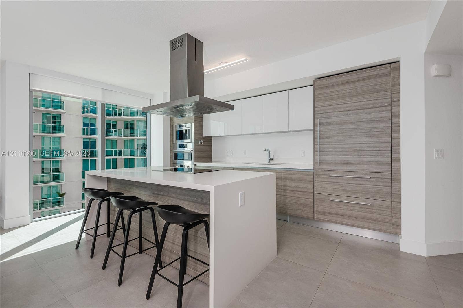Modern kitchen featuring an island with a waterfall-edge countertop, panel-ready refrigerator, stainless steel ceiling-mounted range hood, built-in double wall ovens, and two-tone flat-panel cabinetry