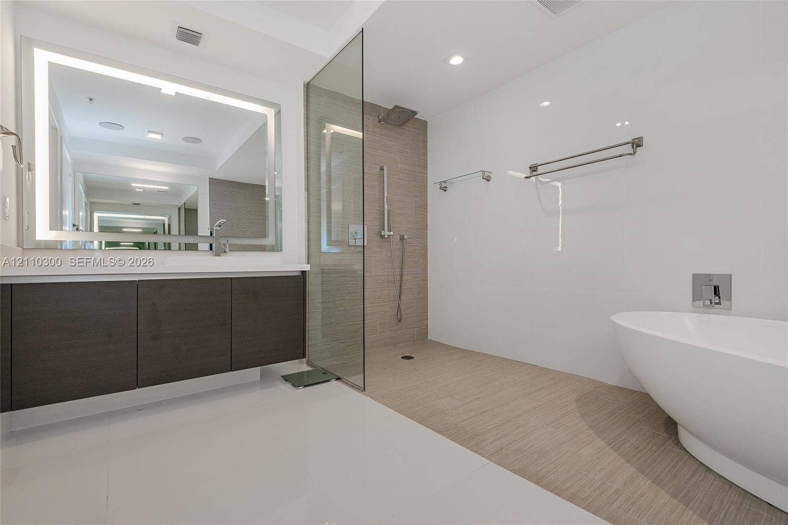 Master Bathroom Floating vanity with dark wood-finish cabinetry, integrated LED-backlit mirror, walk-in glass shower, wall-mounted shower hardware, and freestanding soaking tub