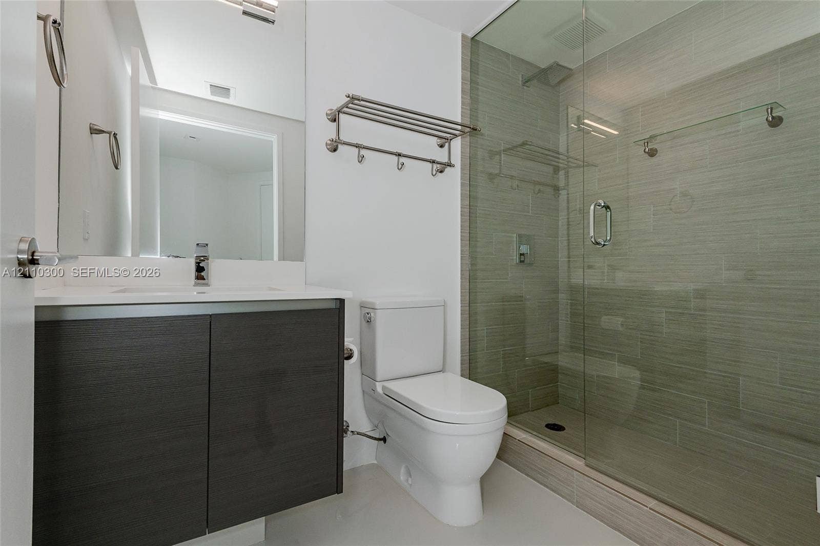 Bathroom featuring a vanity with wood-finish cabinetry and a white countertop, walk-in shower with a glass enclosure and textured tile surround, wall-mounted metal shelving, and a dual-flush toilet