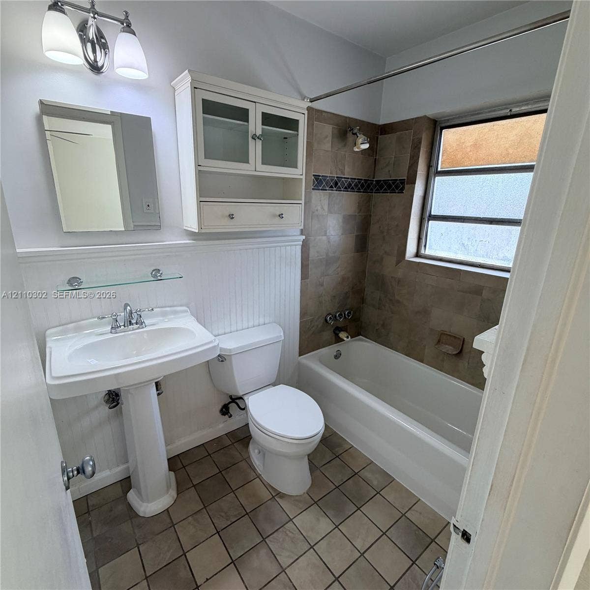 Bathroom featuring a pedestal sink, beadboard wainscoting, wall-mounted storage cabinetry, tiled flooring, and a bathtub with tile surround