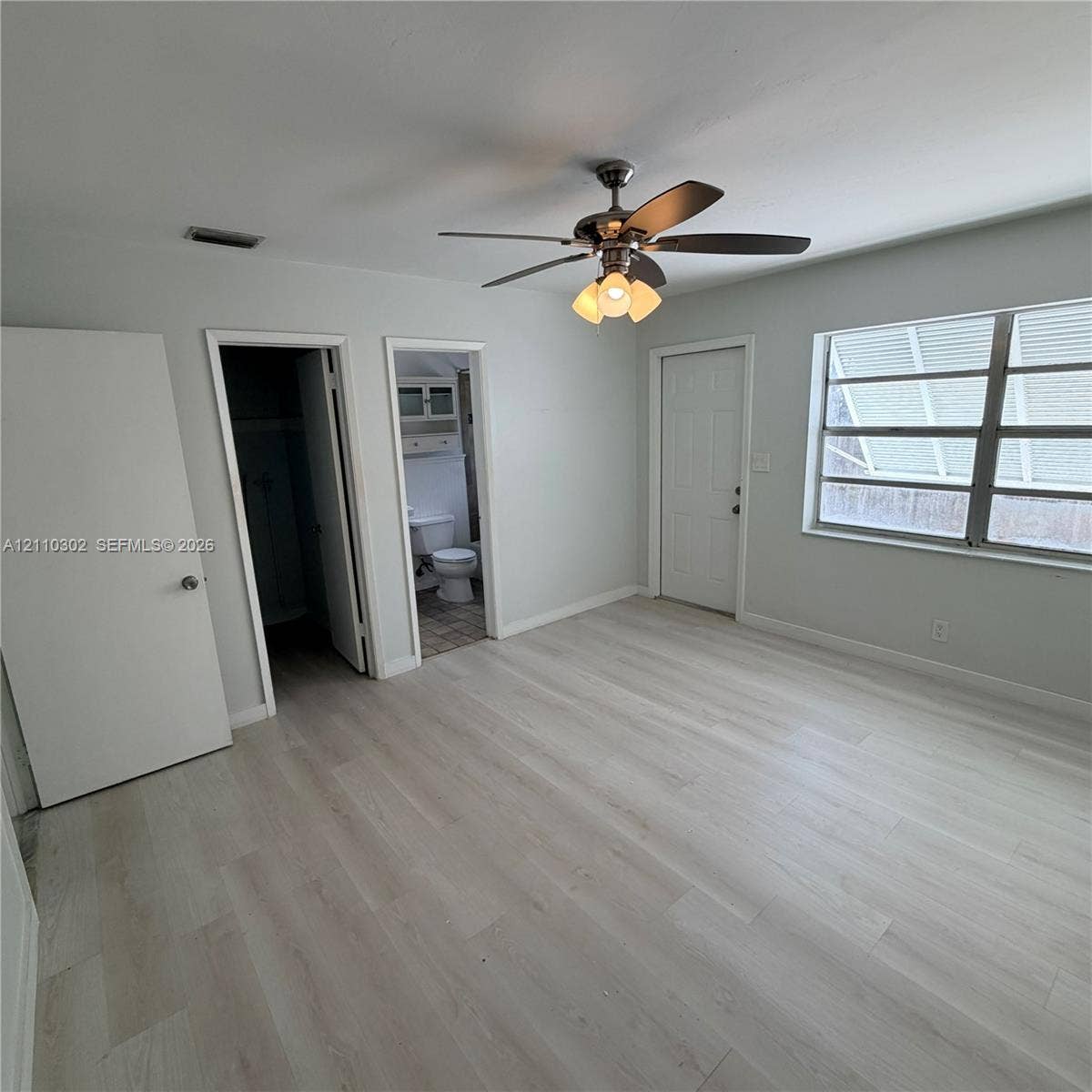 Light-toned wood-finish flooring, central ceiling fan with multi-light fixture, and multi-pane window with adjustable louvers