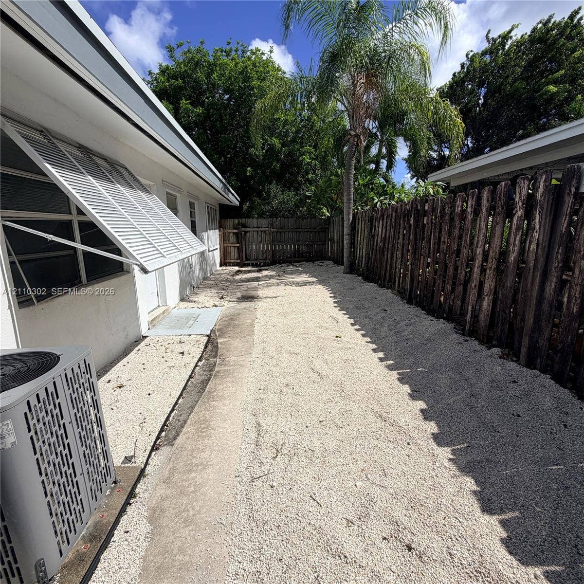 Exterior side yard featuring gravel ground cover, a wood-slat privacy fence, metal window awnings, and mature palm tree landscaping