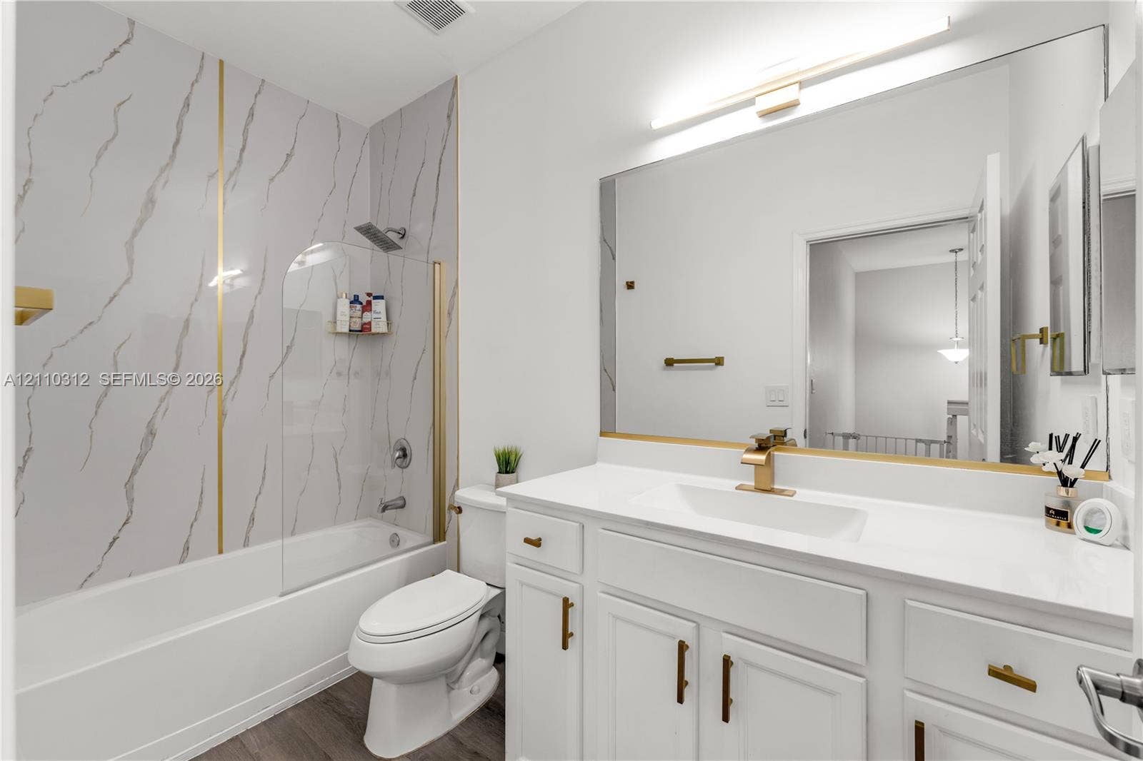 11524 SW 248th Lane, Homestead, FL 33032 Bathroom featuring a white vanity with gold-finish hardware, a large frameless mirror, a shower-tub combination with gold-accented wall panels, a glass panel screen, and wood-finish flooring