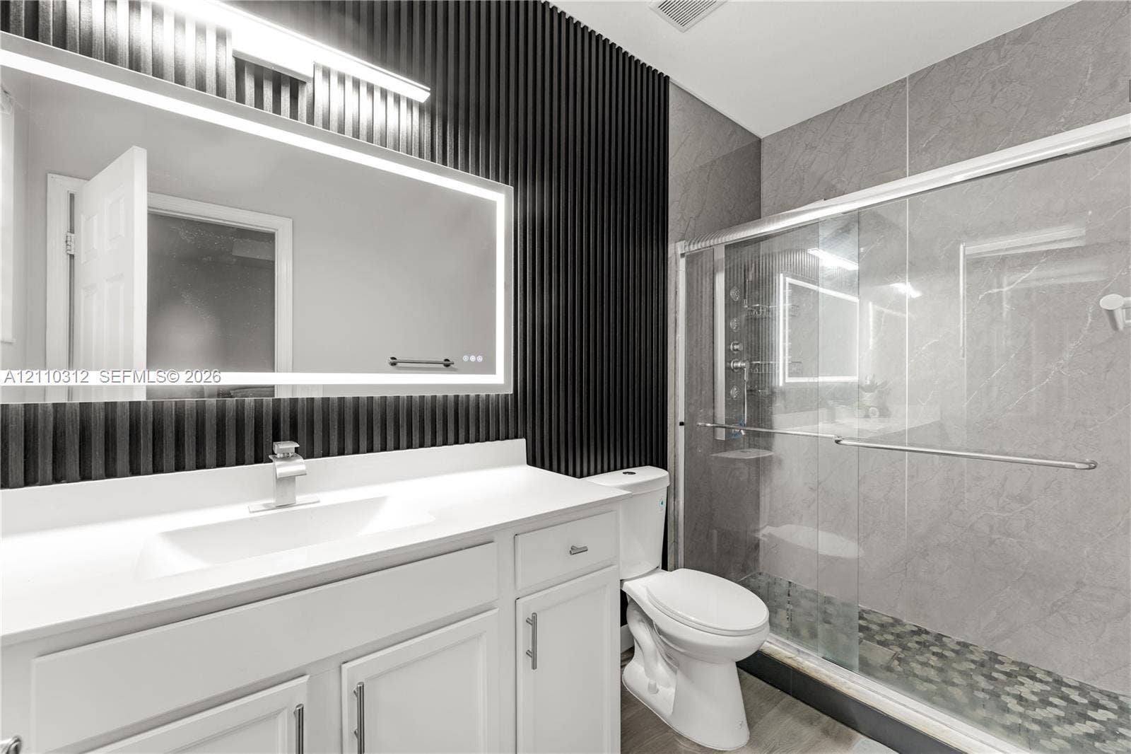 11524 SW 248th Lane, Homestead, FL 33032 Bathroom featuring a white vanity with a rectangular sink, wall-mounted backlit mirror, black vertical slat accent wall, and a walk-in shower with grey tile surround and stone-style flooring