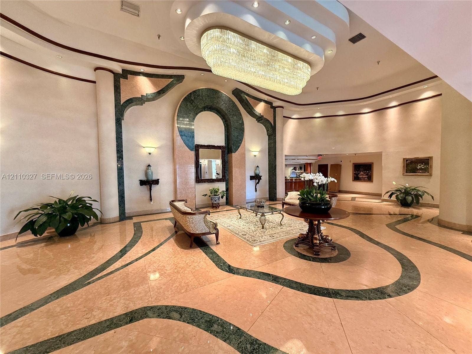 Grand foyer featuring polished stone flooring with intricate inlaid stone accents, a multi-tiered recessed ceiling, a large-scale crystal chandelier, and architectural wall detailing with stone arches