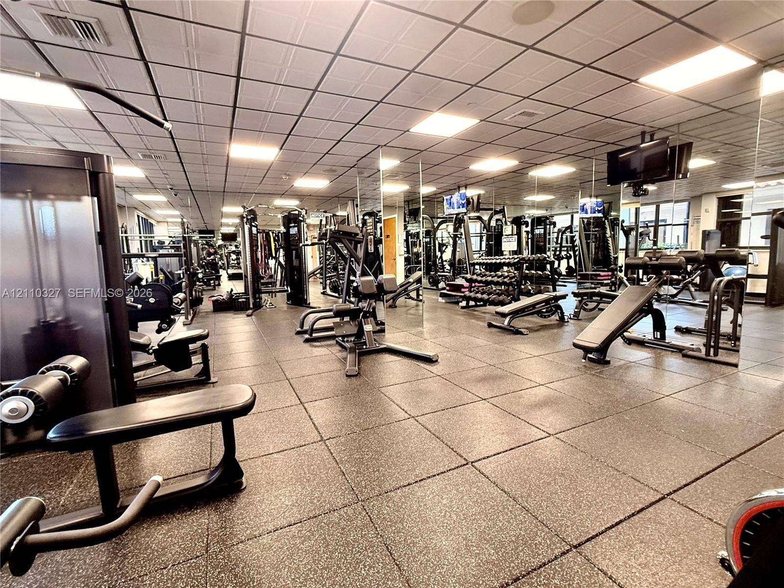 Fitness center featuring grid-pattern acoustic ceiling tiles, recessed lighting, wall-to-wall mirrored surfaces, and high-impact rubber flooring