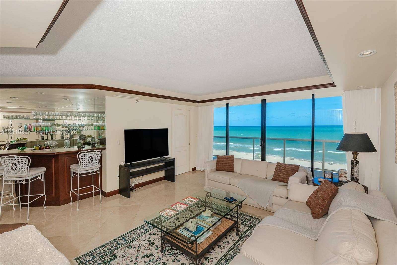 Living area featuring gorgeous ocean views, a custom wood-finished wet bar, stone tile flooring, crown molding, recessed lighting, and expansive floor-to-ceiling glass panel doors opening to a balcony