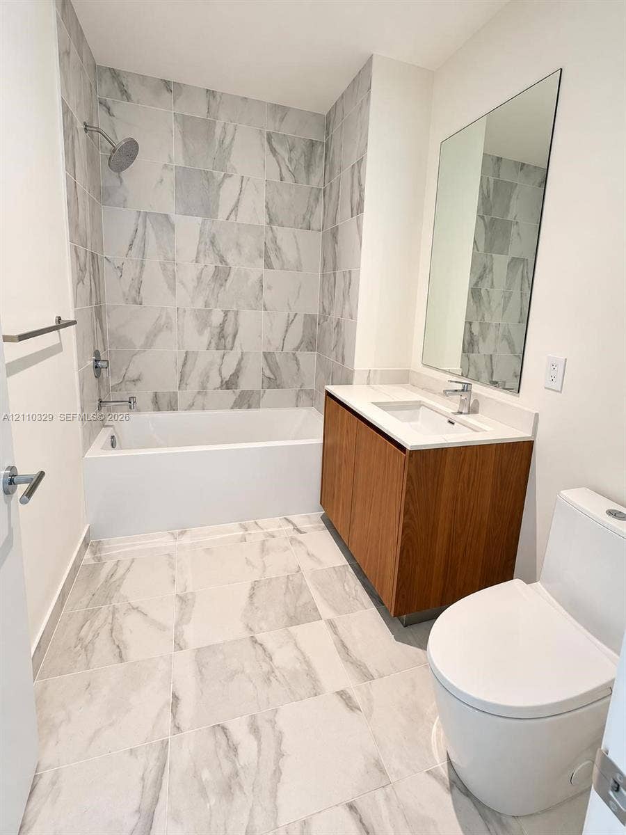 Bathroom featuring large-format tile flooring, wood-finish vanity with a rectangular sink, wall-mounted mirror, and a tile-surround soaking tub with a shower fixture